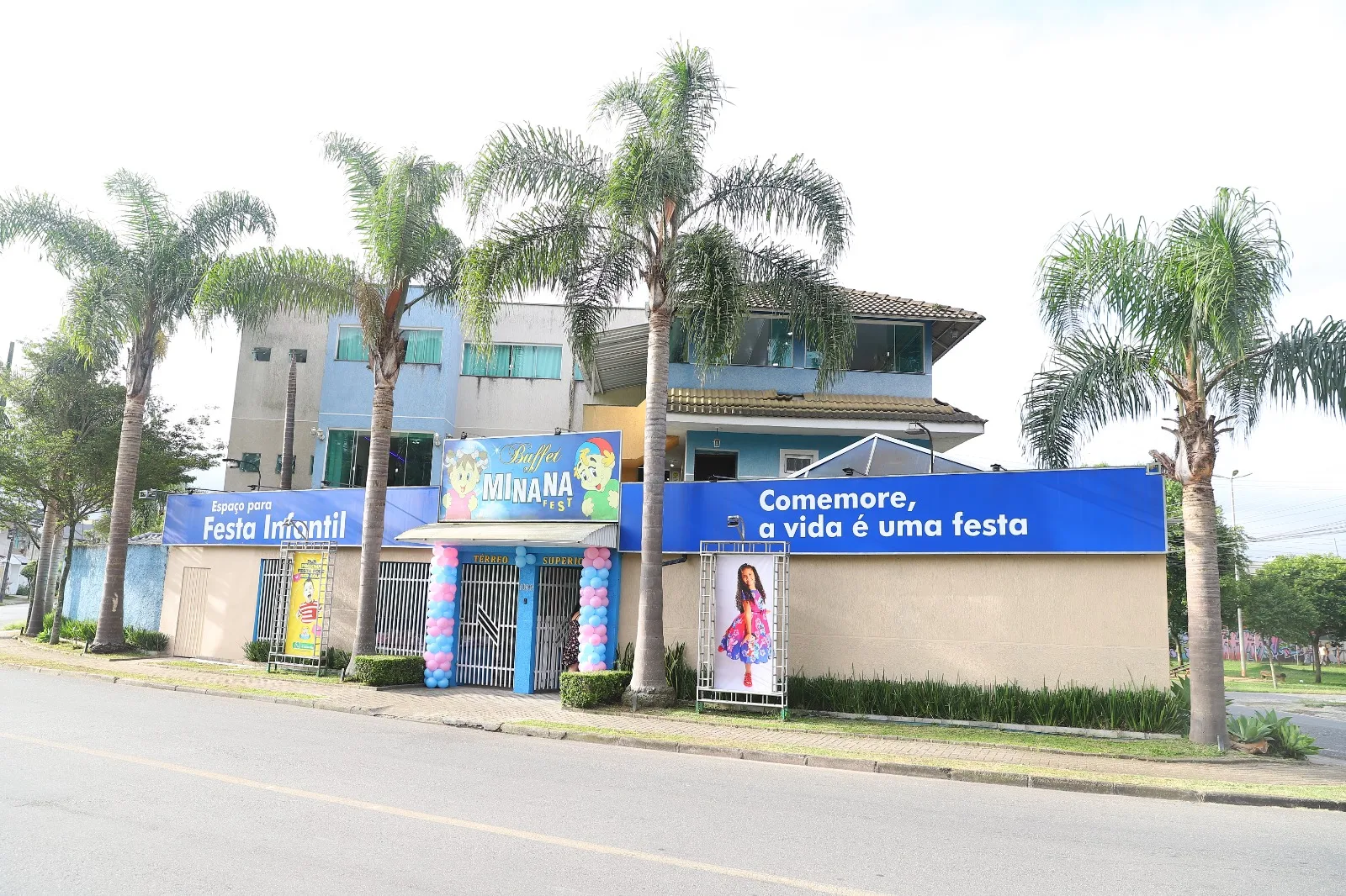 Fachada real do MinanaFest com prédio de dois andares, palmeiras, painel azul 'Comemore, a vida é uma festa' e balões na entrada