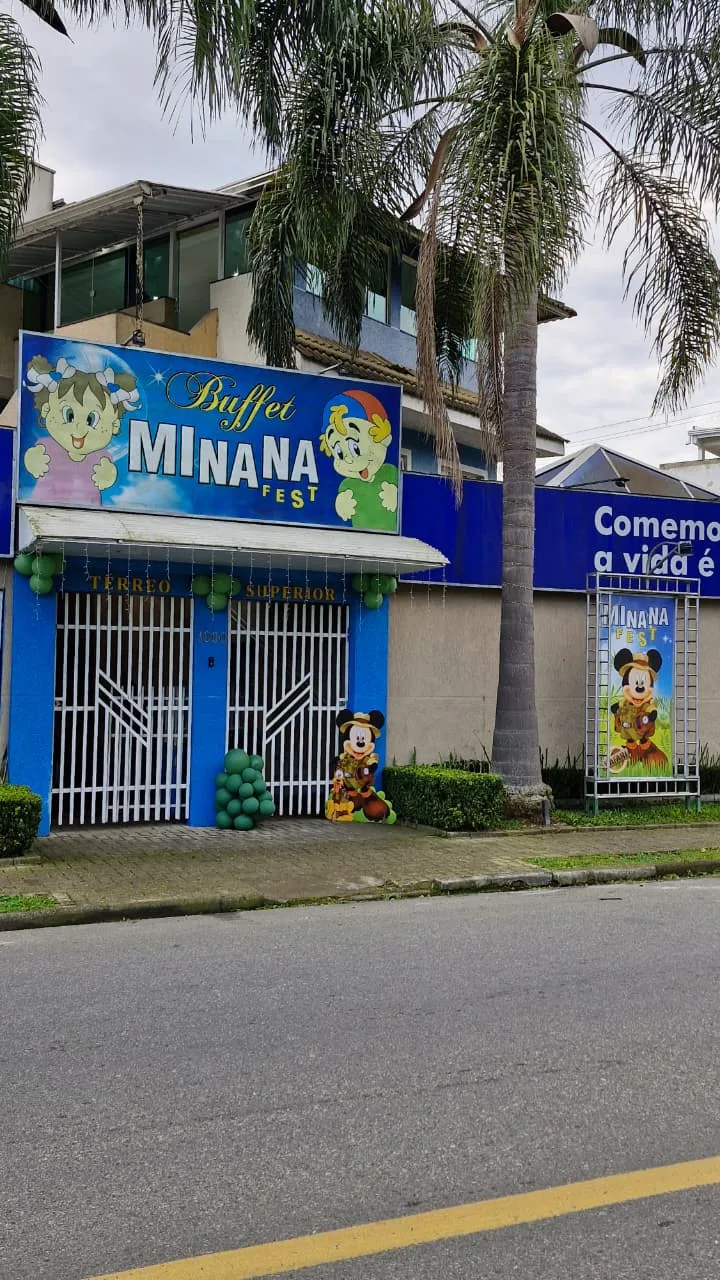 Fachada do MinanaFest em enquadramento vertical mostrando o prédio, palmeiras, painel azul e entrada com balões